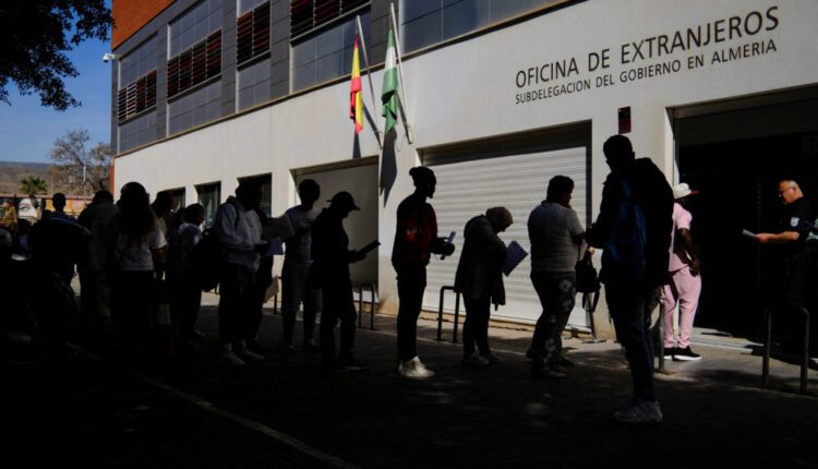 People line up outside a government immigration office in Almería, silhouetted against the building facade with flags nearby.