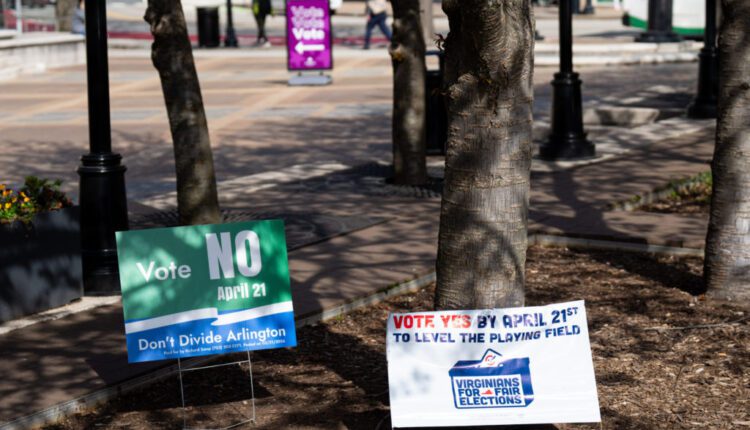 Election signs in a park: a green 'Vote NO' sign and a white 'Vote Yes by April 21st' sign with Virginians for Fair Elections logo, trees in foreground